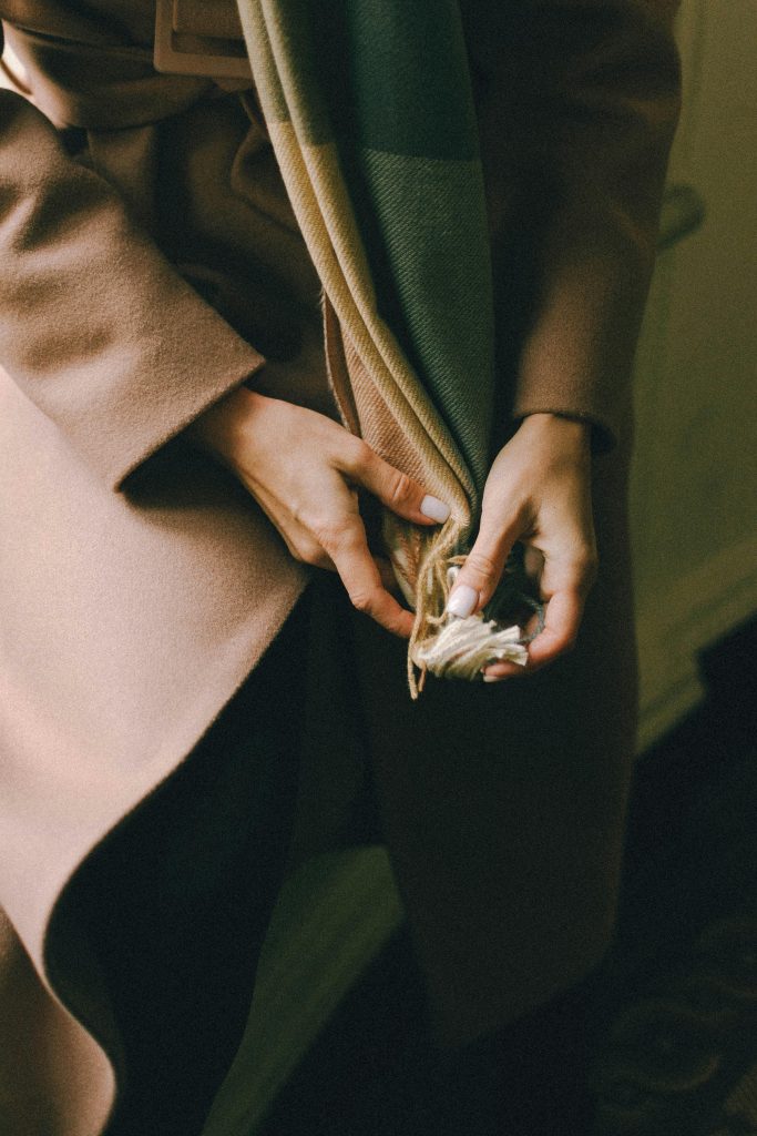 Hands adjusting a warm, neutral-colored scarf paired with a beige coat, creating a cozy and stylish autumn look