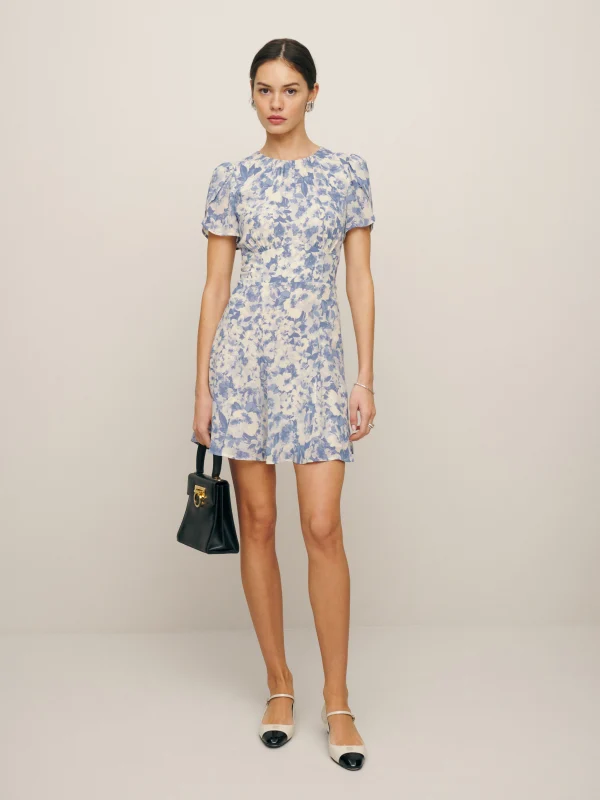 Woman wearing a blue and white floral print mini dress with puff sleeves, carrying a small black handbag and wearing white flats with black toe caps for a fresh and elegant style.