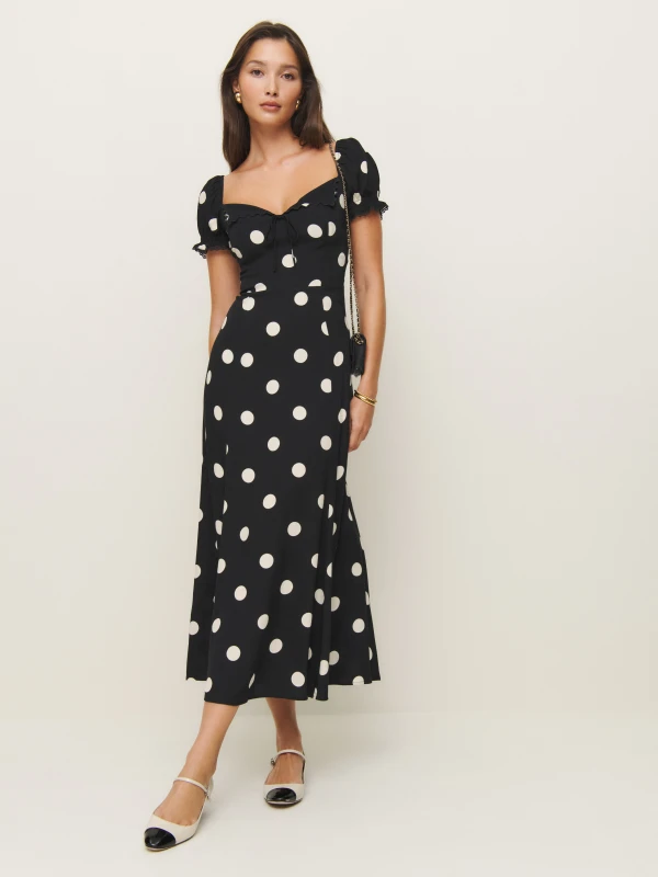 Woman in a black midi dress with white polka dots, puff sleeves, and a sweetheart neckline, paired with white flats featuring black toe caps for a playful yet sophisticated look.