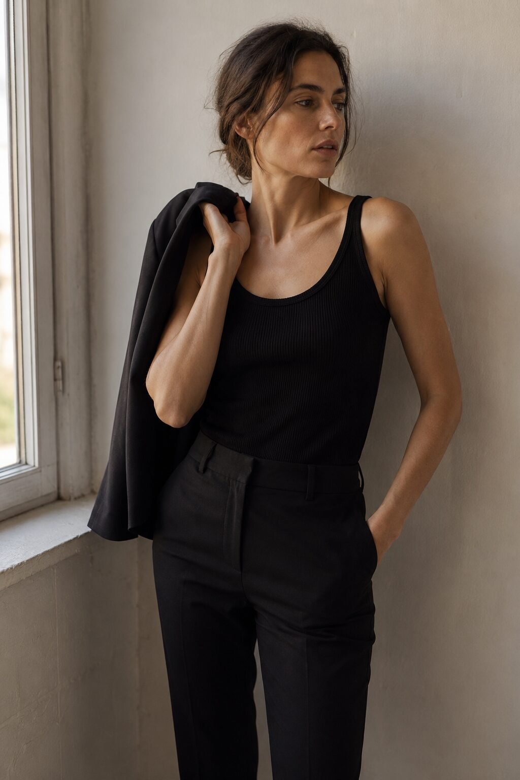 woman in a 90s minimalist outfit wearing a black ribbed tank and tapered cigarette trouser with a blazer carried over one shoulder