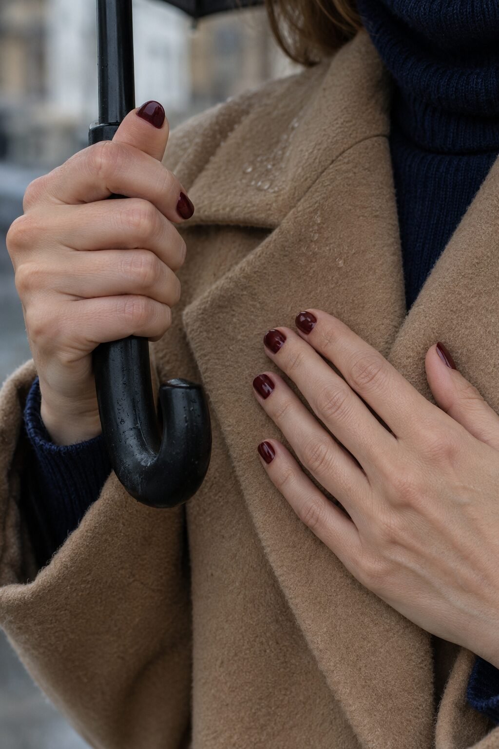 close-up of short glossy oxblood red nails against a camel wool coat, one hand holding an umbrella handle