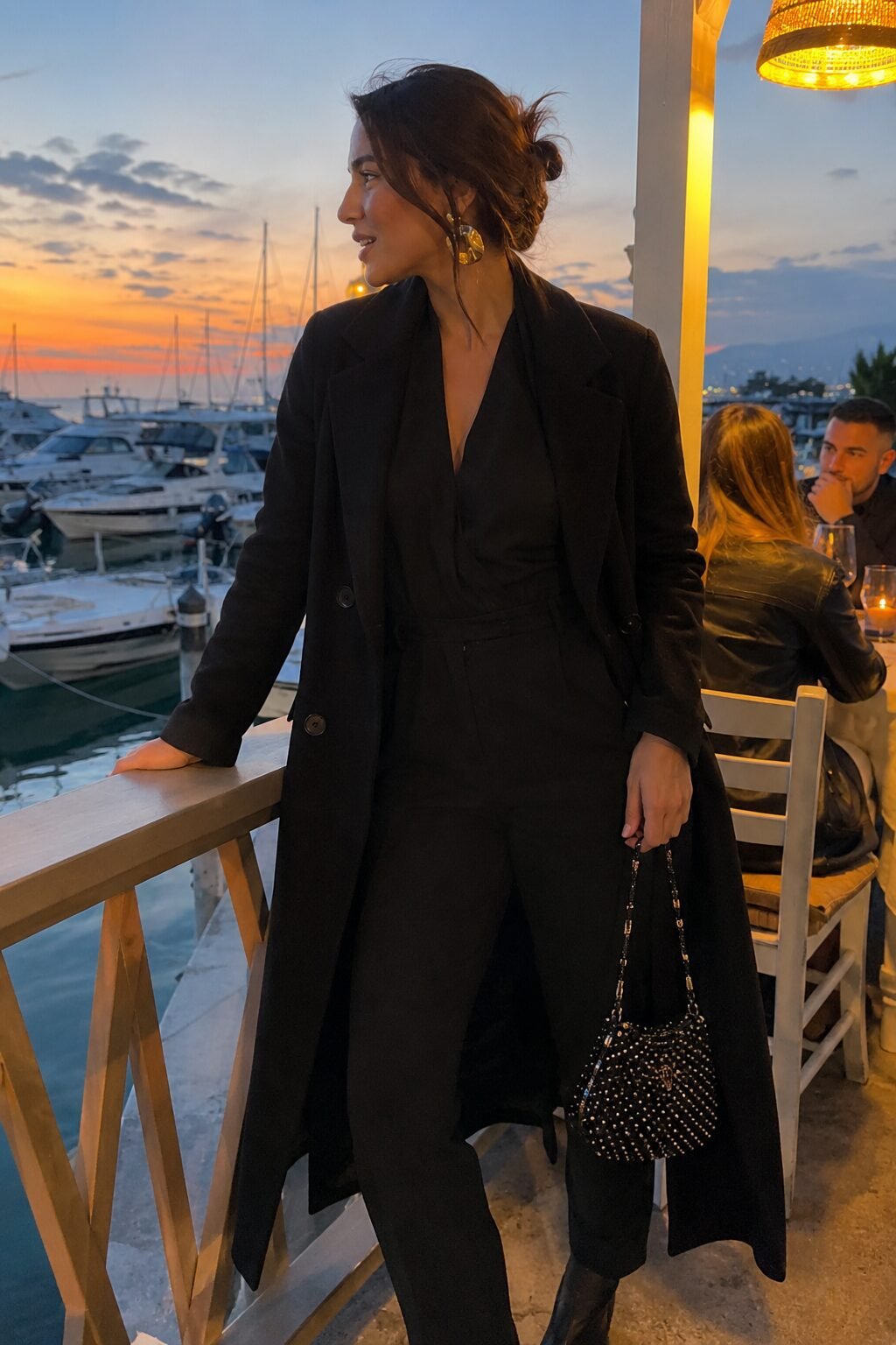 how to style a black trench coat with a jumpsuit: unbelted black trench over a sleek black jumpsuit with a metallic earring for date night