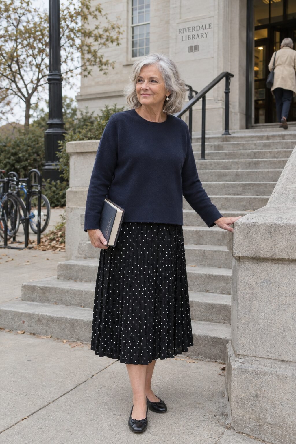 how to style a tunic for women in their 70s: navy crewneck tunic over a black pleated midi skirt with pearl earrings