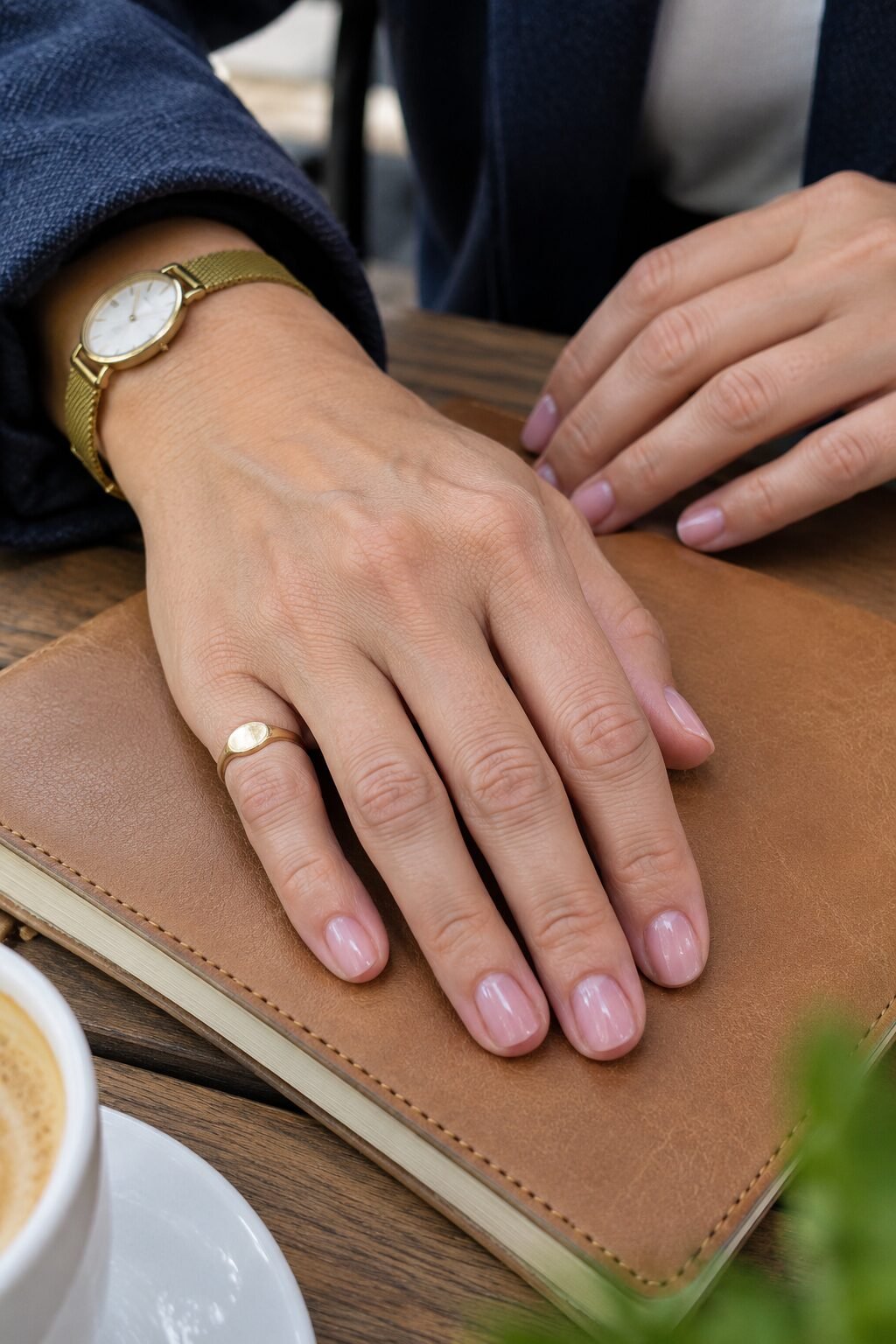 close-up of a hand with sheer pink nails, a gold signet ring and a slim gold watch resting on a leather notebook