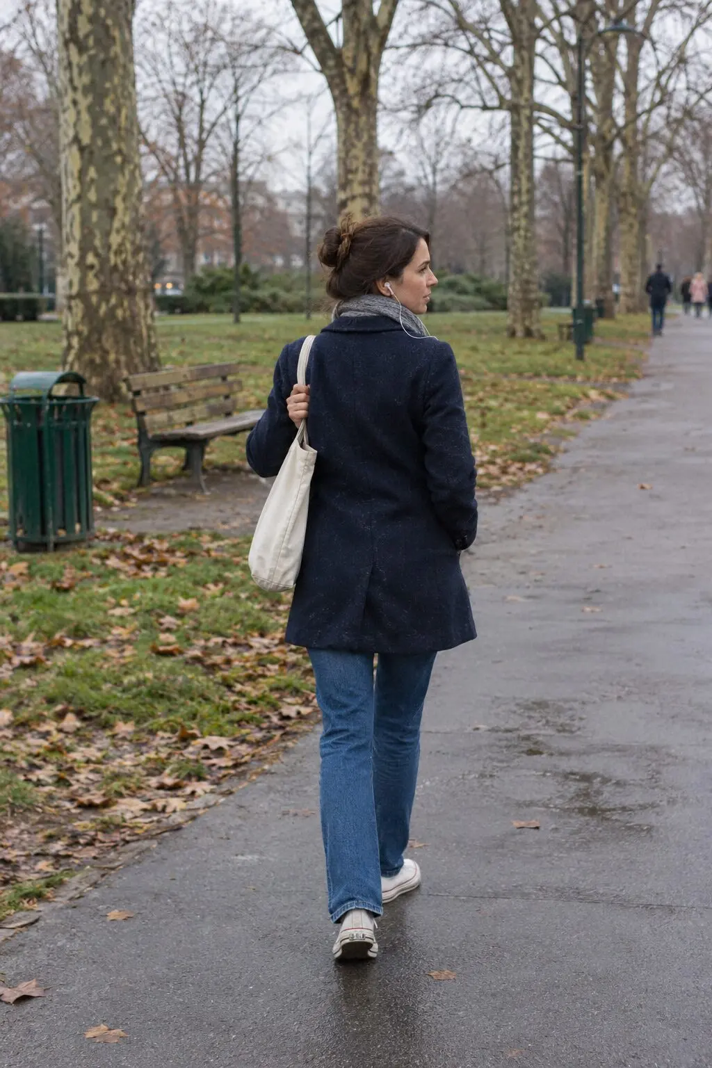 A woman in a navy wool coat and jeans walking away along a quiet park path listening to music on wired earbuds