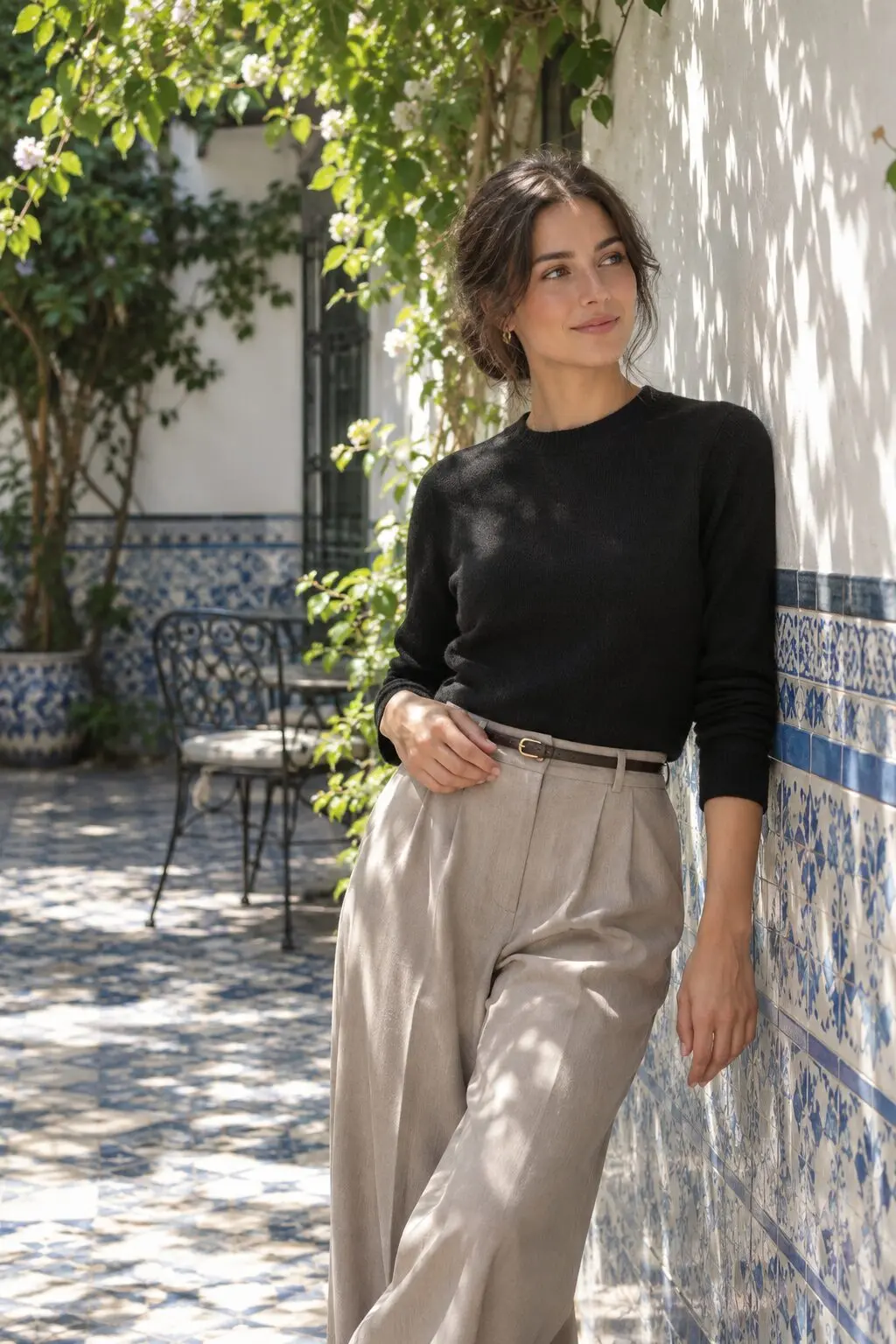 a woman in a black cashmere crewneck and taupe wide-leg trouser leaning against a blue-and-white tiled courtyard wall