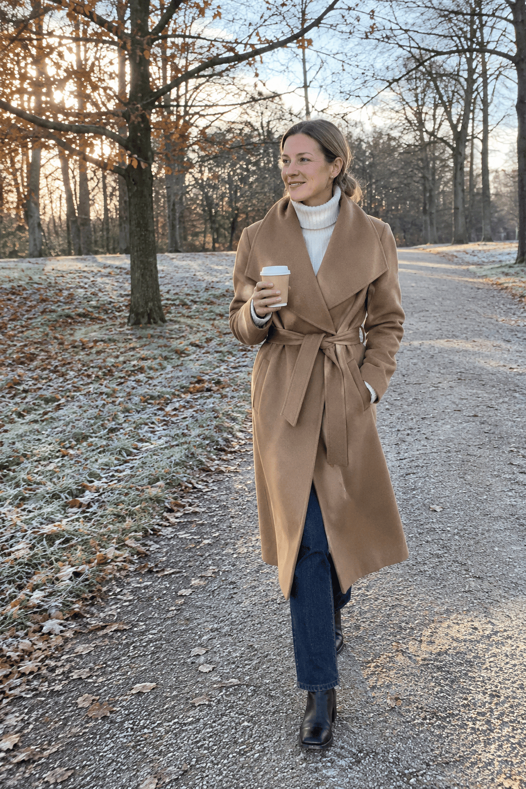 Thanksgiving morning walk outfit: caramel mid-calf wrap coat, white cashmere turtleneck, dark wash jeans and black ankle boots on an autumn path
