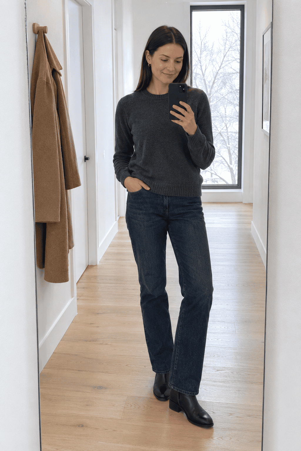 elegant casual winter outfit: woman in a dark gray merino crewneck sweater and straight indigo jeans in a bright hallway