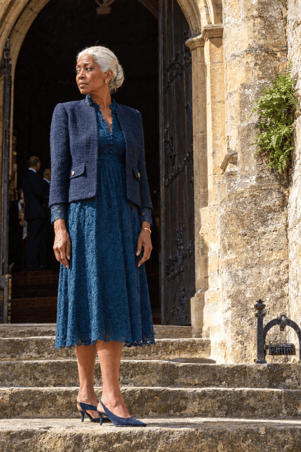elegant church wedding guest outfit for a woman in her 70s: deep petrol blue long-sleeve lace tea dress, navy tweed collarless jacket and navy kitten slingbacks
