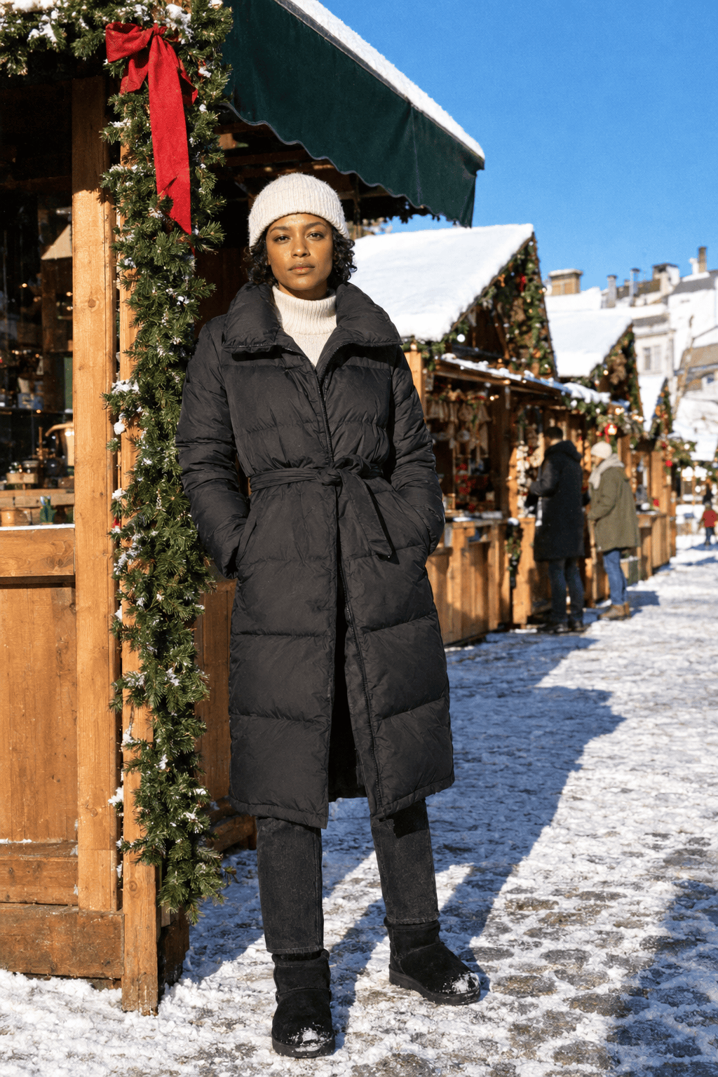 snowy christmas market outfit with a long black belted down puffer coat, cream ribbed wool sweater, black jeans and black shearling-lined boots