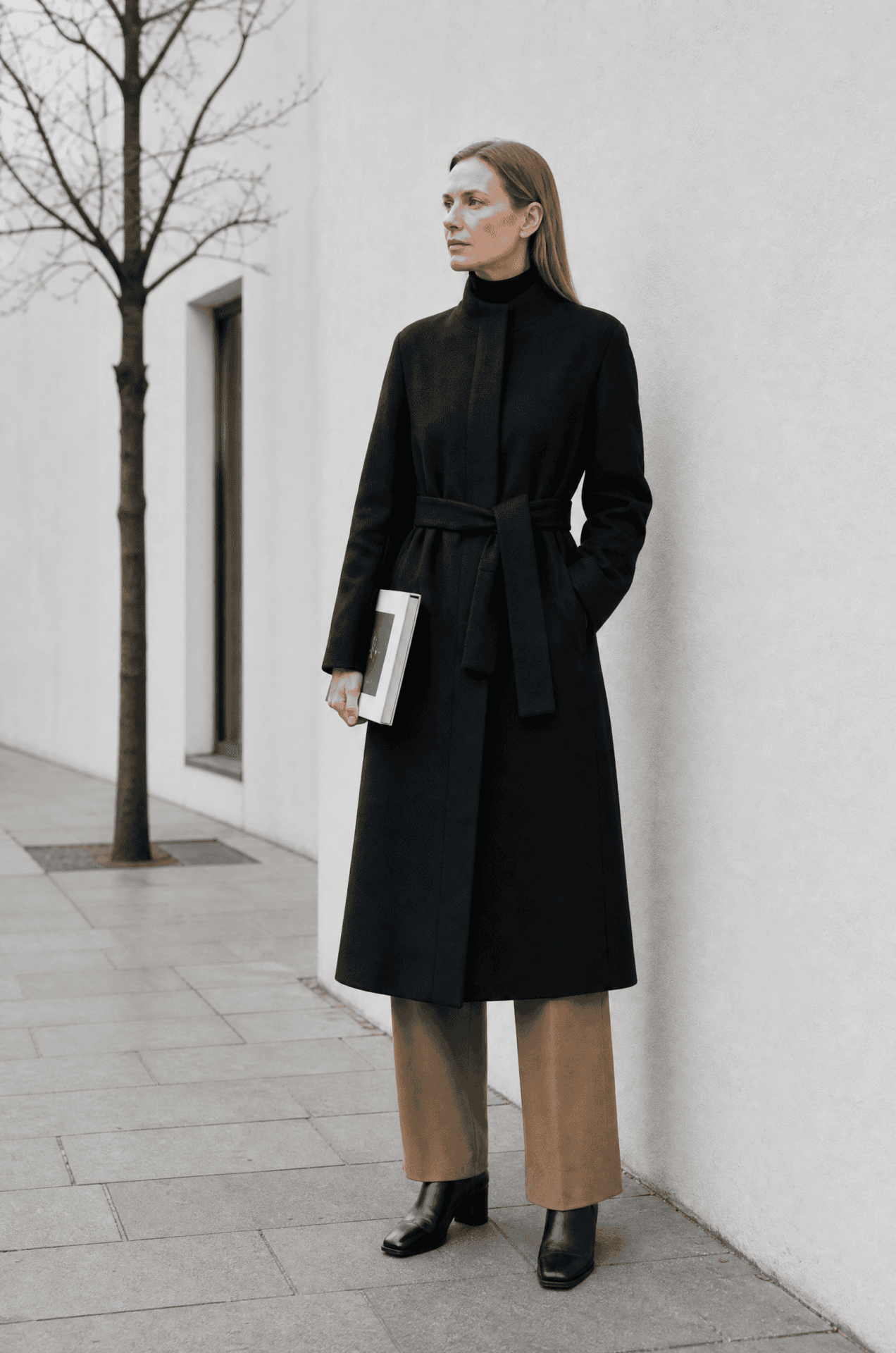 how to wear a long black coat with a black turtleneck: belted black wool coat, black fine-rib turtleneck at the neckline, camel trousers and black ankle boots