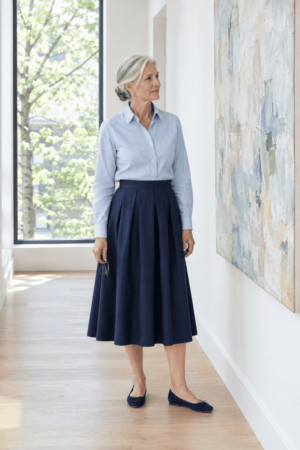 elegant spring outfit for women over 60: light blue striped poplin shirt tucked into a navy pleated mid-calf skirt with navy suede ballet flats