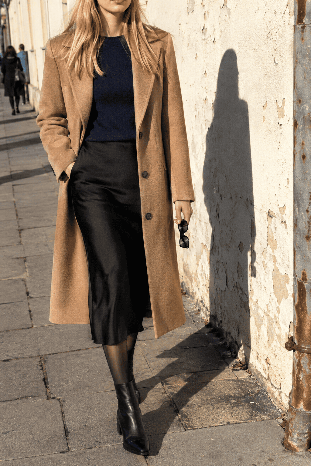 capsule wardrobe evening outfit: camel topcoat over a navy cashmere crewneck and black slip midi skirt with black ankle boots