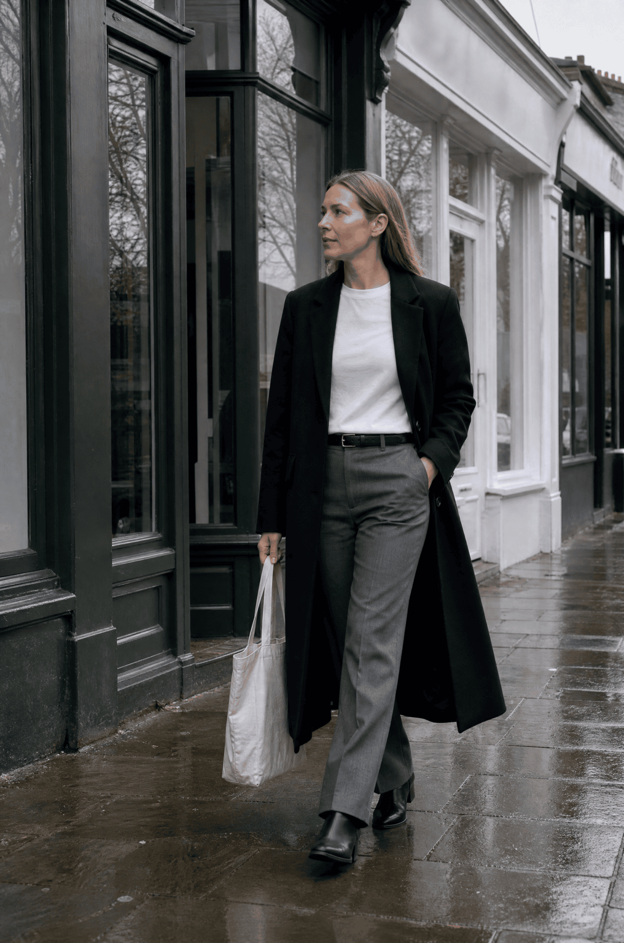 long black coat outfit with a white tee: tailored black coat worn open over a tucked white cotton tee, grey tailored trousers and black ankle boots