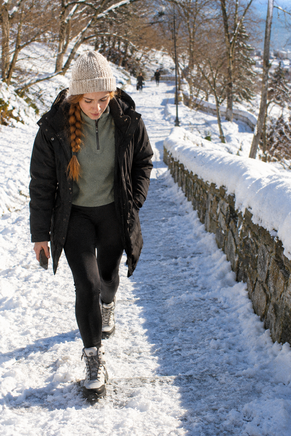 snow outfits for women: black waterproof insulated 3-in-1 winter parka, sage green fleece half-zip, black thermal leggings and white chunky waterproof winter hiking boots on a snowy hill path