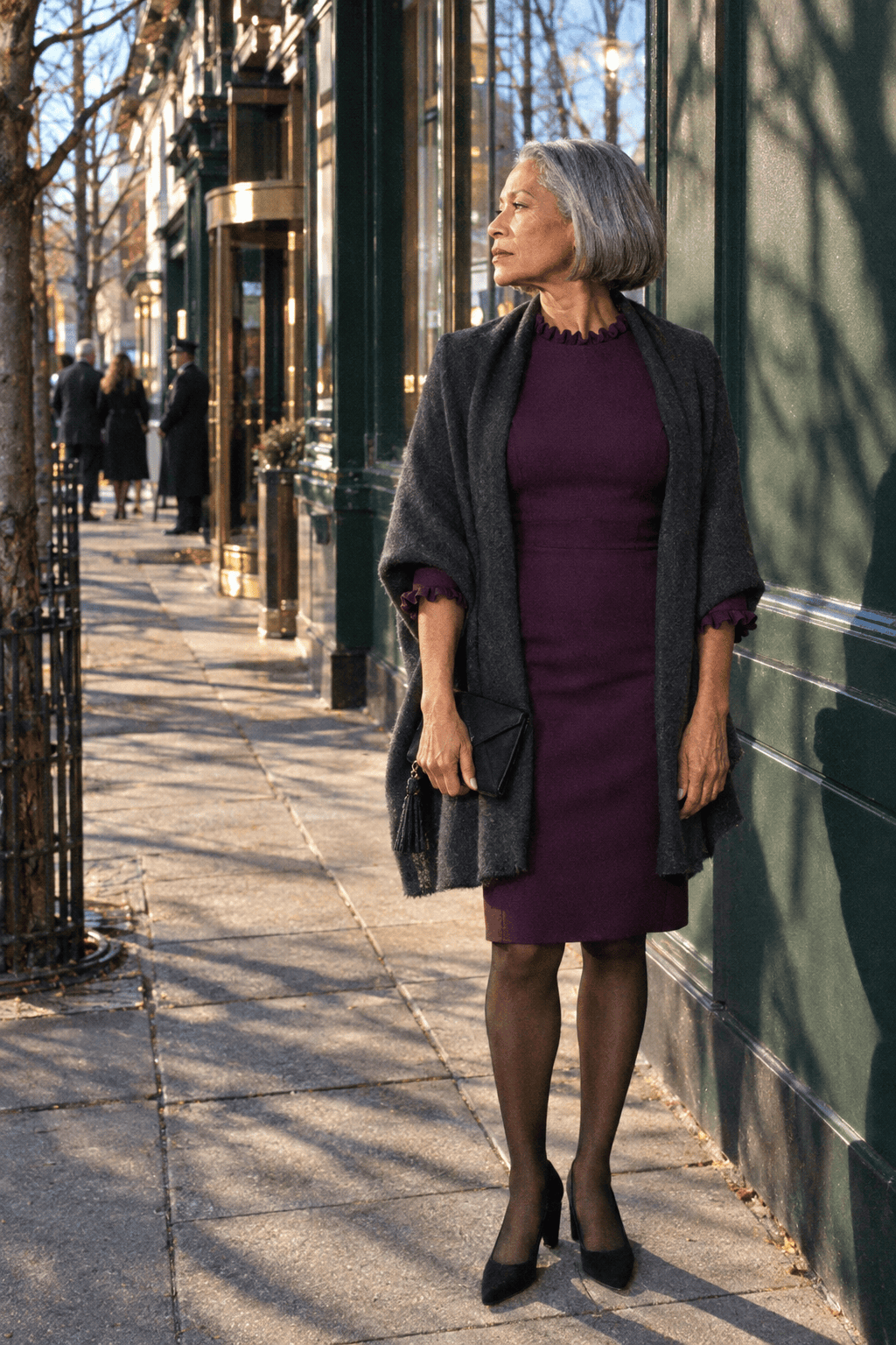 evening hotel wedding guest outfit for a woman in her 70s: deep plum knee-length dress with short ruffle sleeves, charcoal cashmere wrap, black suede block-heel pumps and black satin clutch